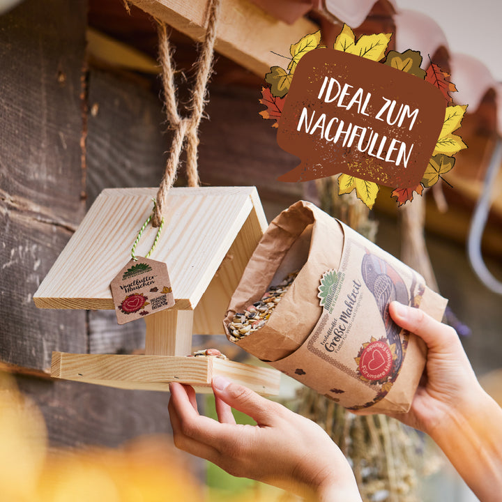 Eine Person füllt ein hölzernes Vogelhaus mit Vogelfutter aus einer Papiertüte; im Bild steht „Ideal zum Nachfüllen“.