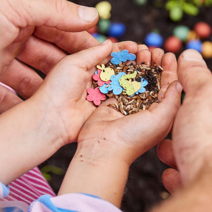 Kinderhände halten bunte Samen in Tier- und Blumenform über Erde.
