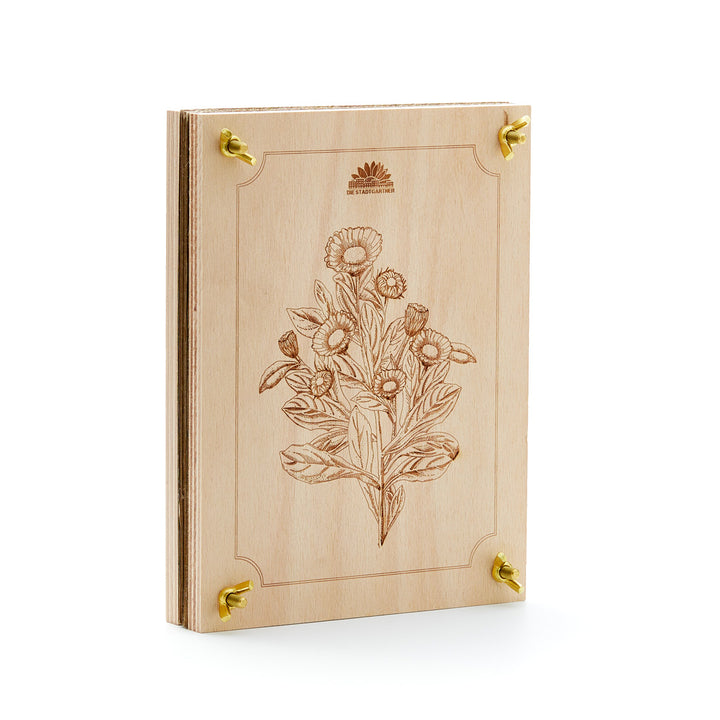 Holzbuch mit floralem Gravurmuster und goldenen Metallecken, stehend auf weißem Hintergrund.