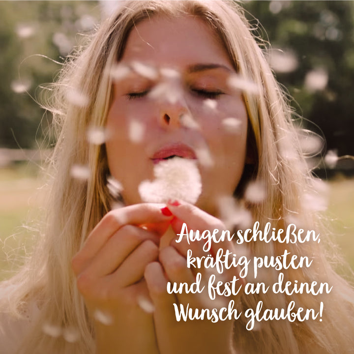 Junge Frau mit geschlossenen Augen pustet eine Pusteblume, während Samen durch die Luft fliegen; Text: „Augen schließen, kräftig pusten und fest an deinen Wunsch glauben!“