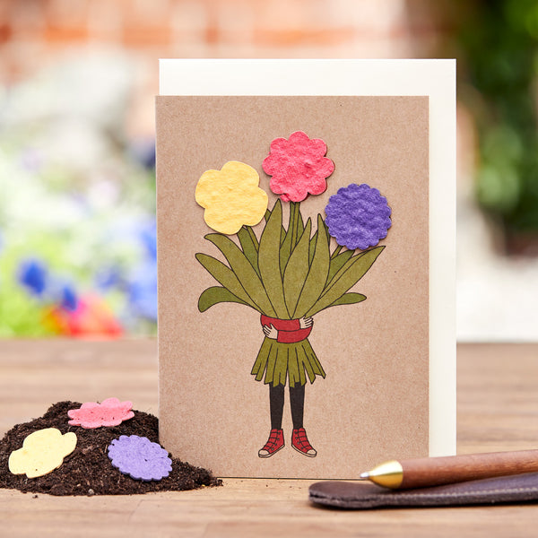 Handgefertigte Grußkarte mit gezeichneten Beinen in roten Schuhen, die einen Strauß bunter Papierblumen halten, auf einem Holztisch mit einem Stift daneben.