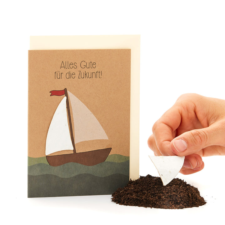 Grußkarte mit Segelboot-Motiv und der Aufschrift „Alles Gute für die Zukunft!“, daneben eine Hand, die ein Segel-förmiges Stück Papier in Erde steckt.